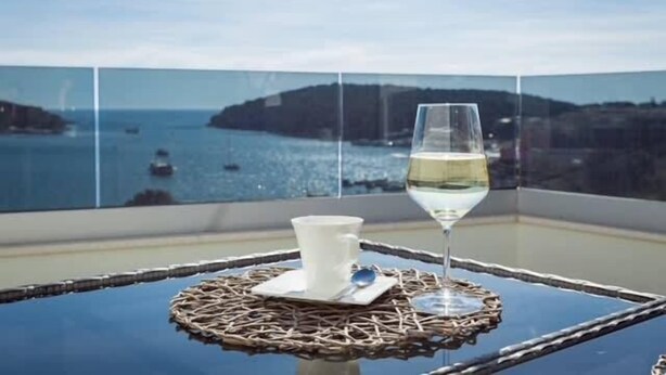 Une table en verre sur laquelle sont posés un gobelet blanc et un verre à vin, avec une vue sur la côte, les bateaux et les collines.