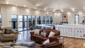 Amplia sala de estar con sofá de cuero marrón, un sillón beige y una mesa de centro blanca. Hay una zona de cocina con encimera blanca y una mesa de comedor. Grandes ventanales ofrecen vistas al exterior.