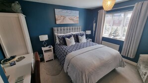 Un dormitorio con cama, mesitas de noche, cómoda, ventana con cortinas y un cuadro con motivos playeros en la pared.
