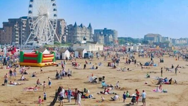 Una playa con una noria al fondo, arena y varias personas recreándose.