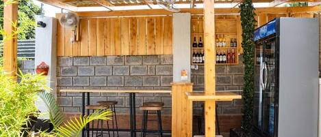 An outdoor bar area with wooden tables and chairs, a stone wall, and a refrigerator.