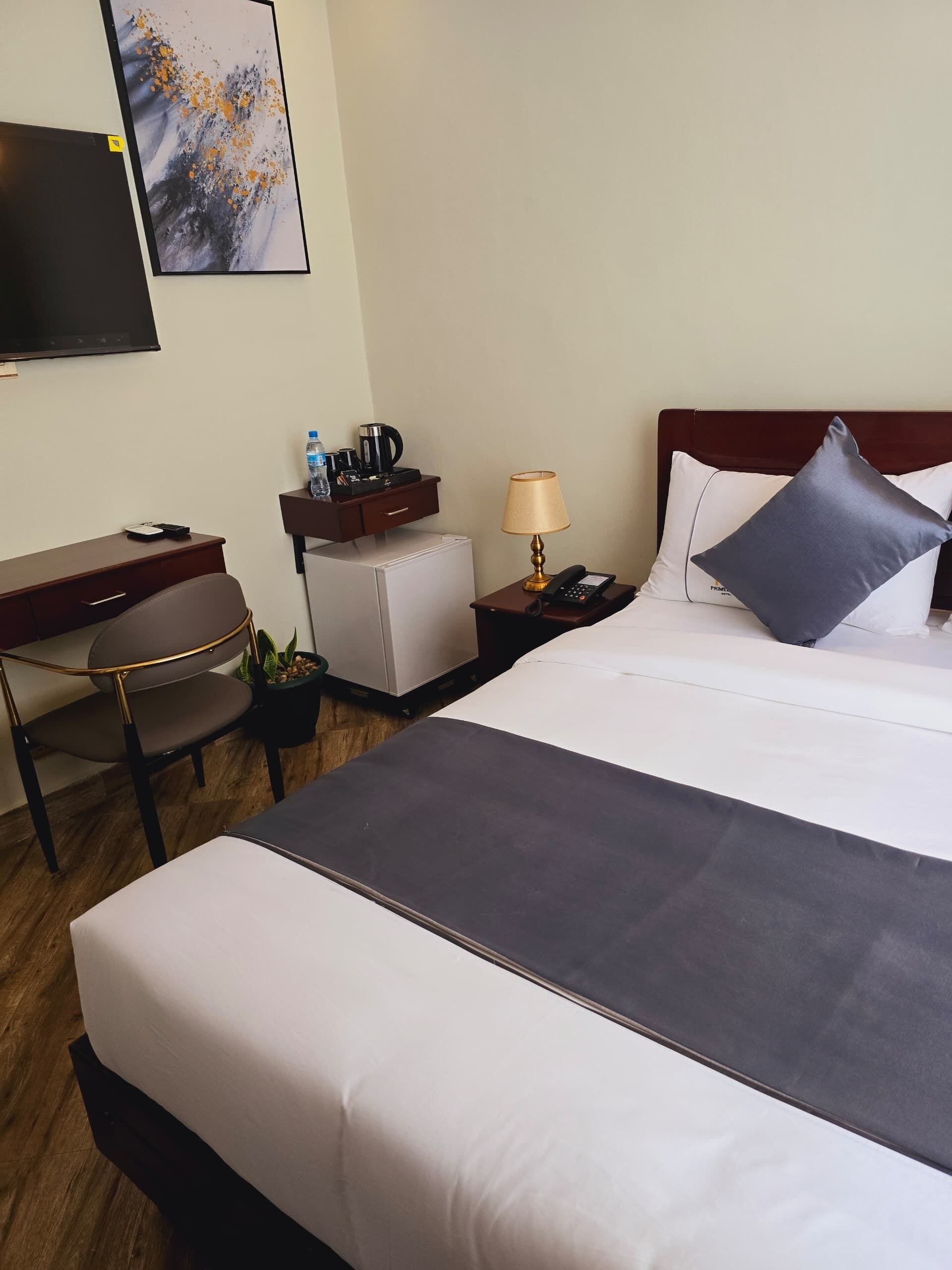 A hotel room with a bed, a desk, a chair, a nightstand, a lamp, and a television on the wall.