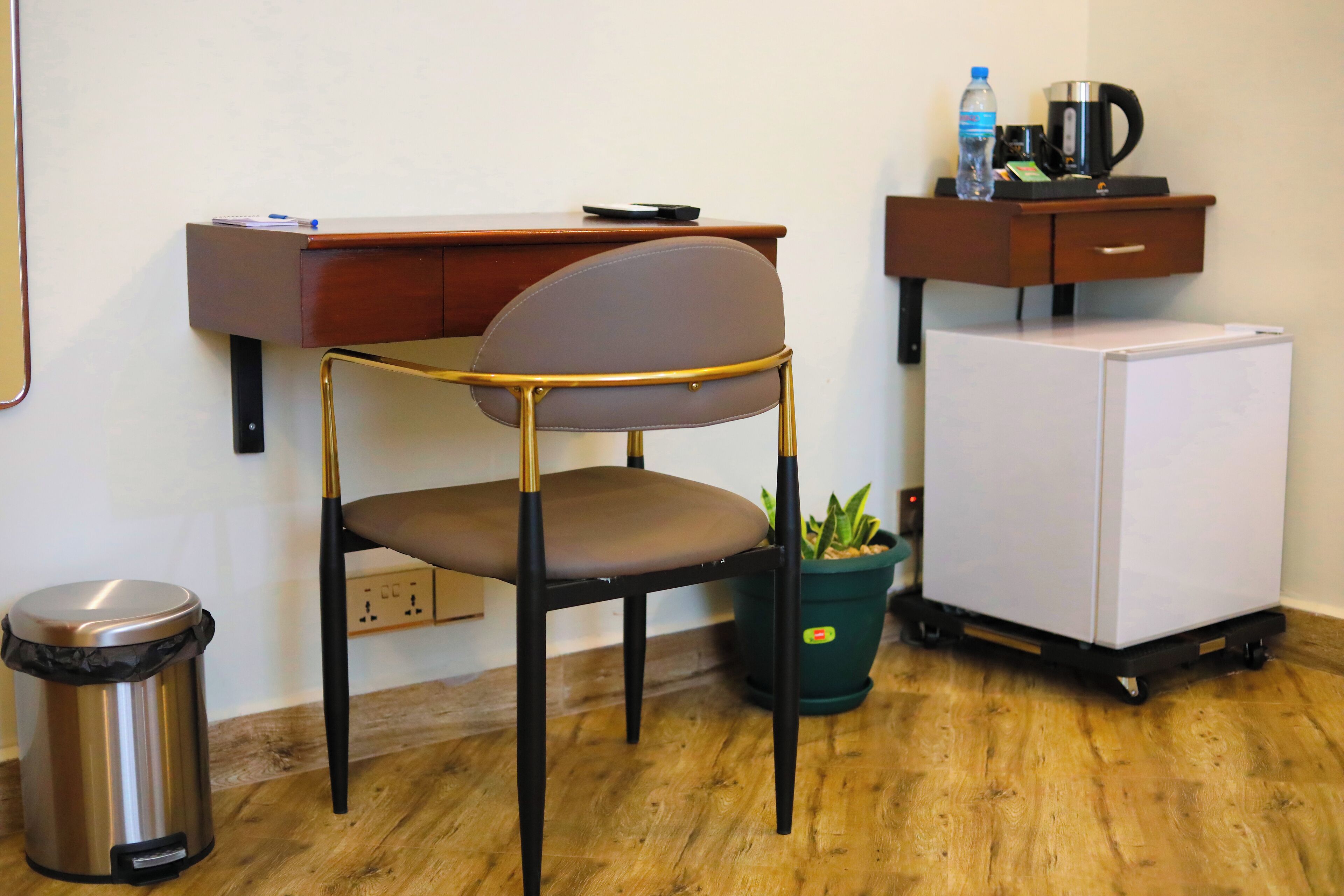 Une petite pièce comprenant un bureau, une chaise, une poubelle, un réfrigérateur et une plante en pot.