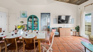 A dining area with a table set for four, a teal cabinet with board games, a television mounted on the wall, and a view of the beach through the window.