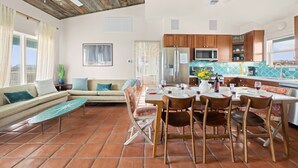 A spacious living area with a sofa, dining table, and chairs, a kitchen with wooden cabinets and a tiled backsplash, and a ceiling fan.