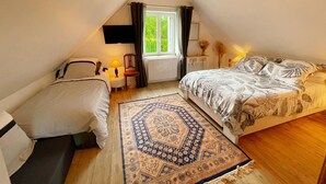 A cozy attic bedroom with a bed, a chair, a small table with a lamp, and a window with curtains. - Charming Cottage with Modern Amenities, Large Garden, and Terrace in Scenic Touraine (Saint-Aubin-le-Dépeint)