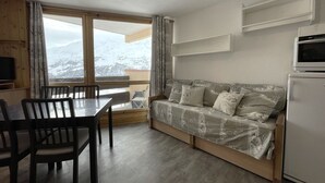 A cozy room with a wooden table and chairs, a sofa with patterned cushions, a small kitchen area with a microwave, and a large window offering a view of a snowy landscape. - Cozy Studio for 3, Ski-in/Ski-out, Balcony, South Exposure, Preyerand, Les Menuires (Les Menuires)
