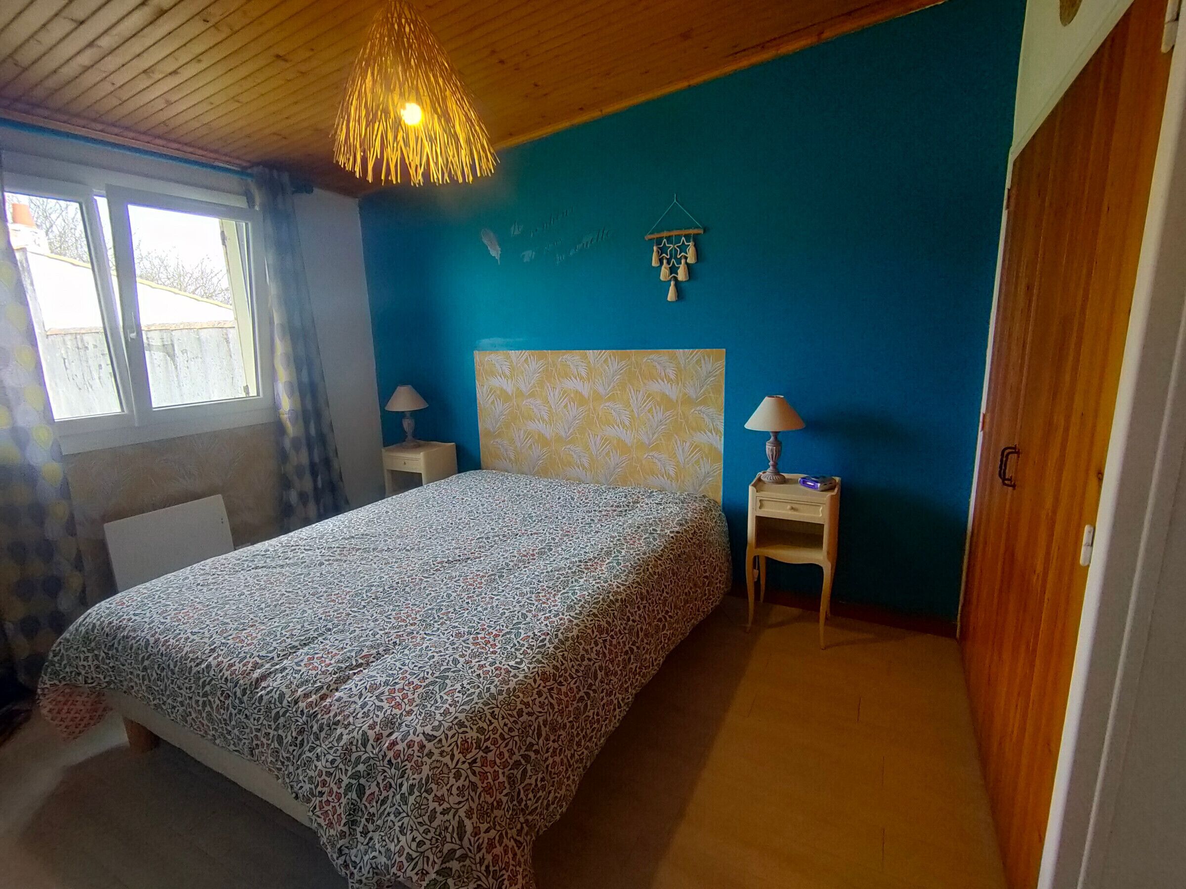 A bedroom with a patterned bedspread, a wooden ceiling, a blue wall, a nightstand with a lamp, and a window with curtains.