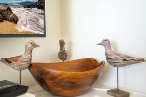 Un tazón de madera con tres esculturas de pájaros sobre pedestales, un televisor mostrando una escena de playa y un dispositivo electrónico negro sobre una superficie blanca.