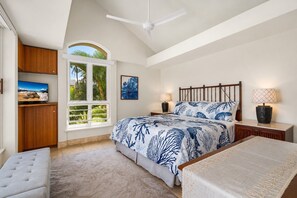 A bedroom with a large bed, a TV, a window with a view of palm trees, a ceiling fan, and a nightstand with a lamp.