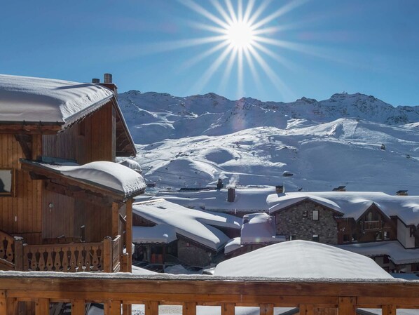 A wooden chalet with a snow-covered roof, set against a backdrop of snow-covered mountains and a clear sky.