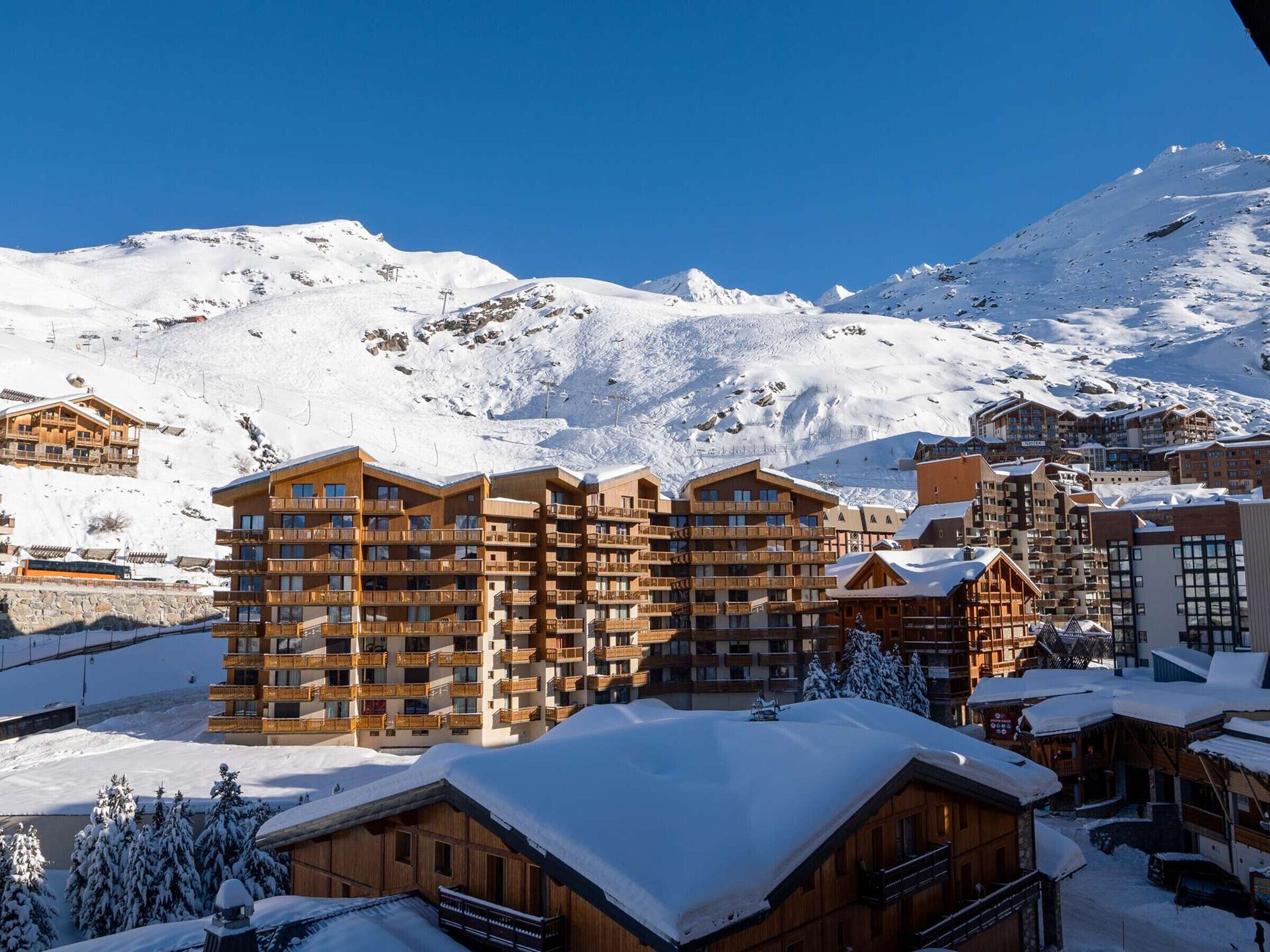 Une station de montagne enneigée, avec des bâtiments en bois à plusieurs étages, des remontées mécaniques et un ciel d’un bleu limpide.