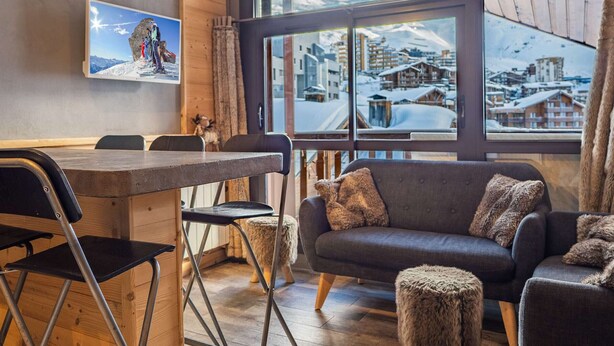 Una habitación acogedora con paredes de madera, un sofá, una mesa de comedor y un amplio ventanal con vistas a una montaña nevada.
