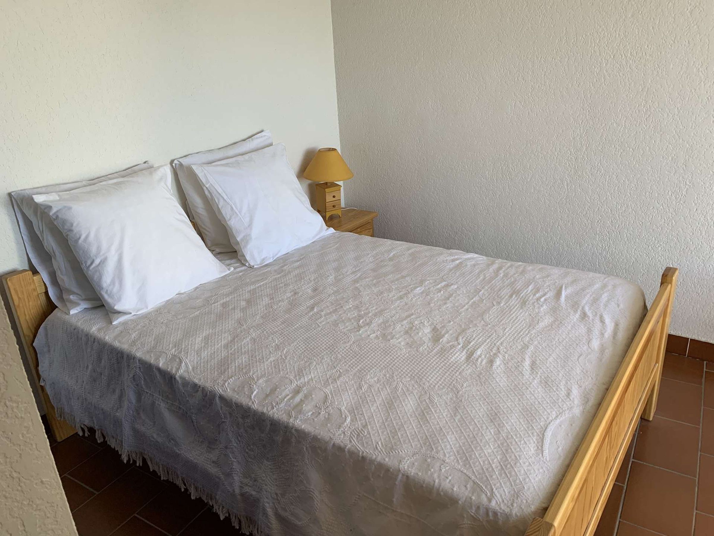 A bed with a white bedspread and two pillows, a wooden nightstand with a lamp, and a plain wall.