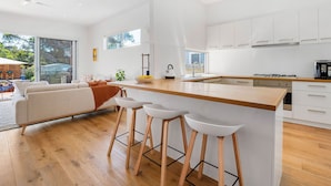 A modern kitchen with white cabinets, a wooden countertop, and a built-in stove. There are three white stools at the island, and a large window provides natural light.