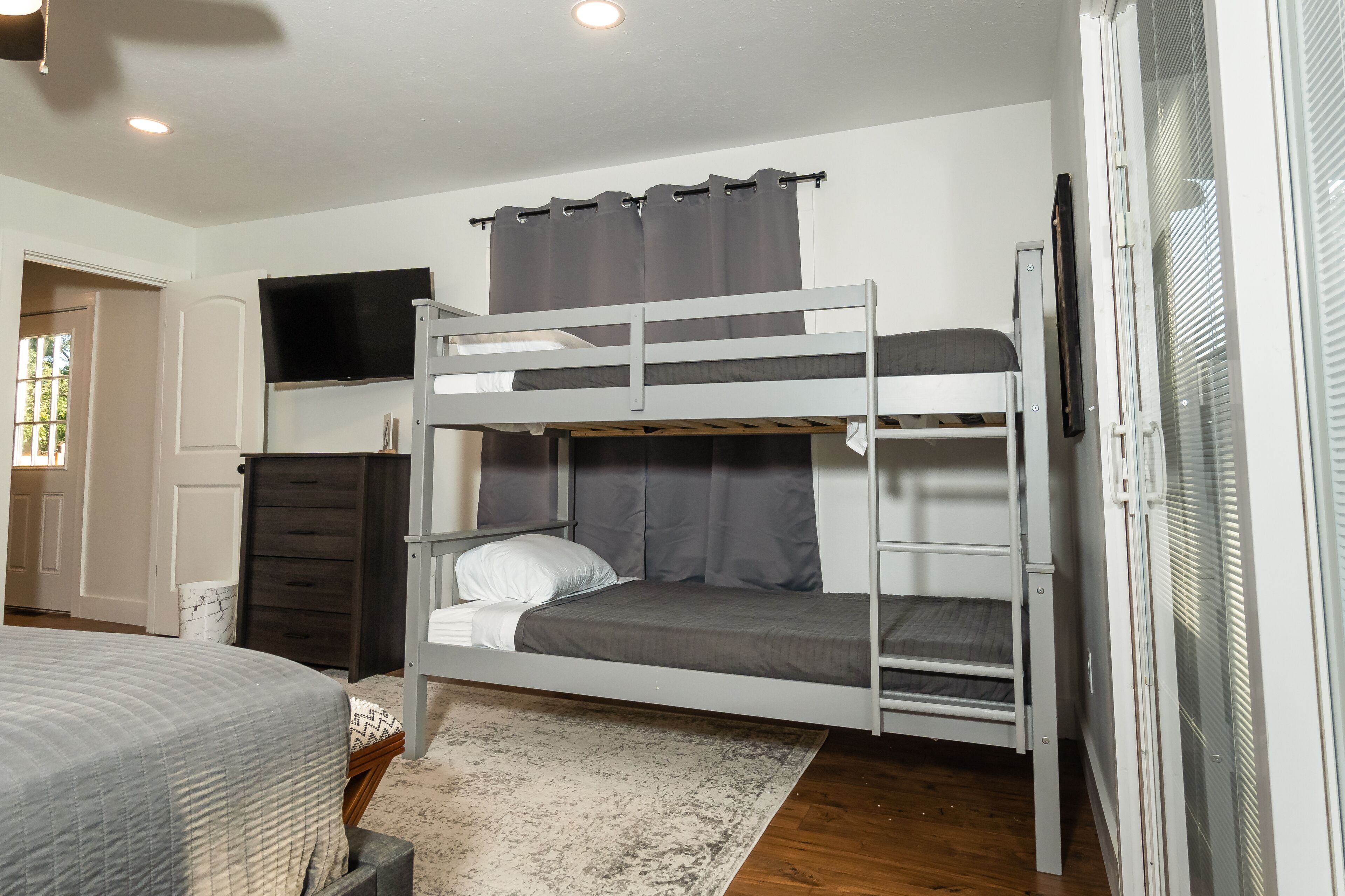 Een slaapkamer met een stapelbed, een televisie boven het bovenste bed, een donkere kledingkast en een raam met zonwering.