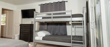 Een slaapkamer met een stapelbed, een televisie boven het bovenste bed, een donkere kledingkast en een raam met zonwering.