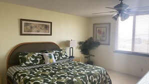 Un dormitorio con cama, ventilador de techo, mesita de noche, lámpara y dos cuadros en la pared.