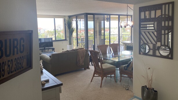 Un salón con una mesa de comedor de vidrio, sillas, un sofá y un televisor. Hay un balcón con vistas a edificios y árboles.