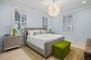 Een slaapkamer met een bed, nachtkastjes, een groene poef en grote ramen met luiken.