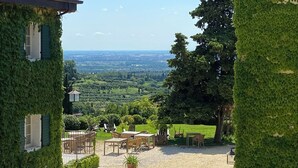 Ein Innenhof mit Steinpflaster, umgeben von mit Efeu bewachsenen Gebäuden. Freiluftmöbel und ein Blick auf die Landschaft sind zu sehen.