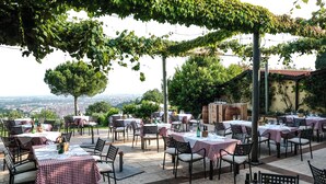 Ein Essbereich im Freien mit Tischen und Stühlen, überdacht von einer mit Wein bewachsenen Pergola, mit Blick auf eine Stadtlandschaft.