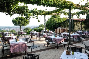 Een buiten eethoek met tafels en stoelen, overdekt door een pergola begroeid met wijnranken, met uitzicht op een stadsbeeld.