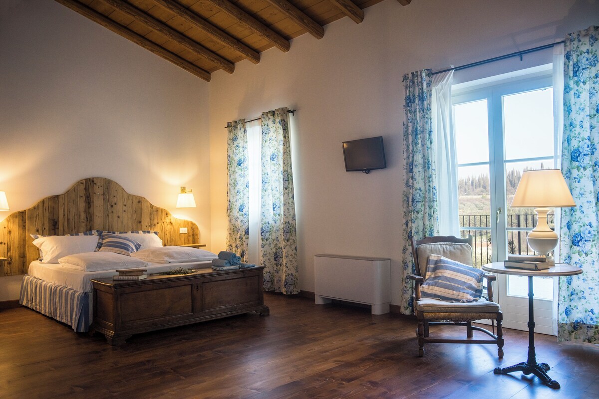 Ein Schlafzimmer mit Bettgestell aus Holz, einem Nachttisch mit Lampe, einem Sessel mit Kissen und einem Fenster mit floralem Vorhang.