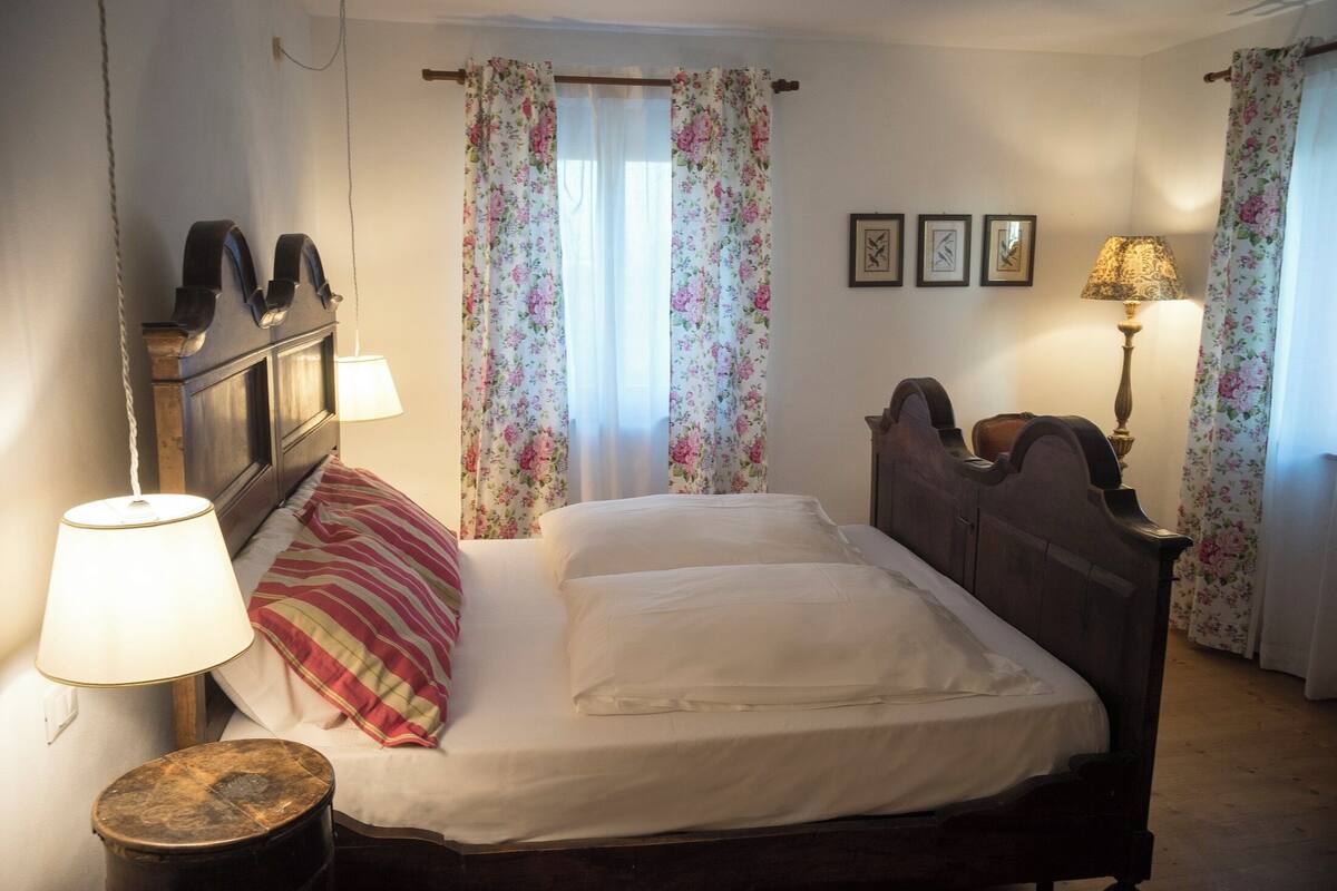 Une chambre à coucher avec un lit, deux lampes de chevet, une tête de lit en bois, des rideaux à motifs floraux et une petite table ronde.