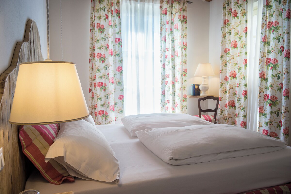 Ein ordentlich bezogenes Bett mit weißen Leinen und gestreiften Kissen, eine Nachttischlampe und blumige Vorhänge.