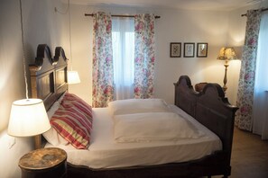 Une chambre à coucher avec un lit, deux lampes de chevet, une tête de lit en bois, des rideaux à motifs floraux et une petite table ronde.