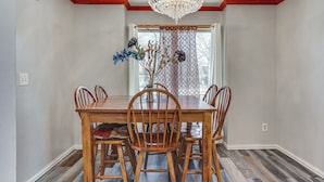 A dining room with a wooden table, four chairs, a chandelier, and a window with a patterned curtain.