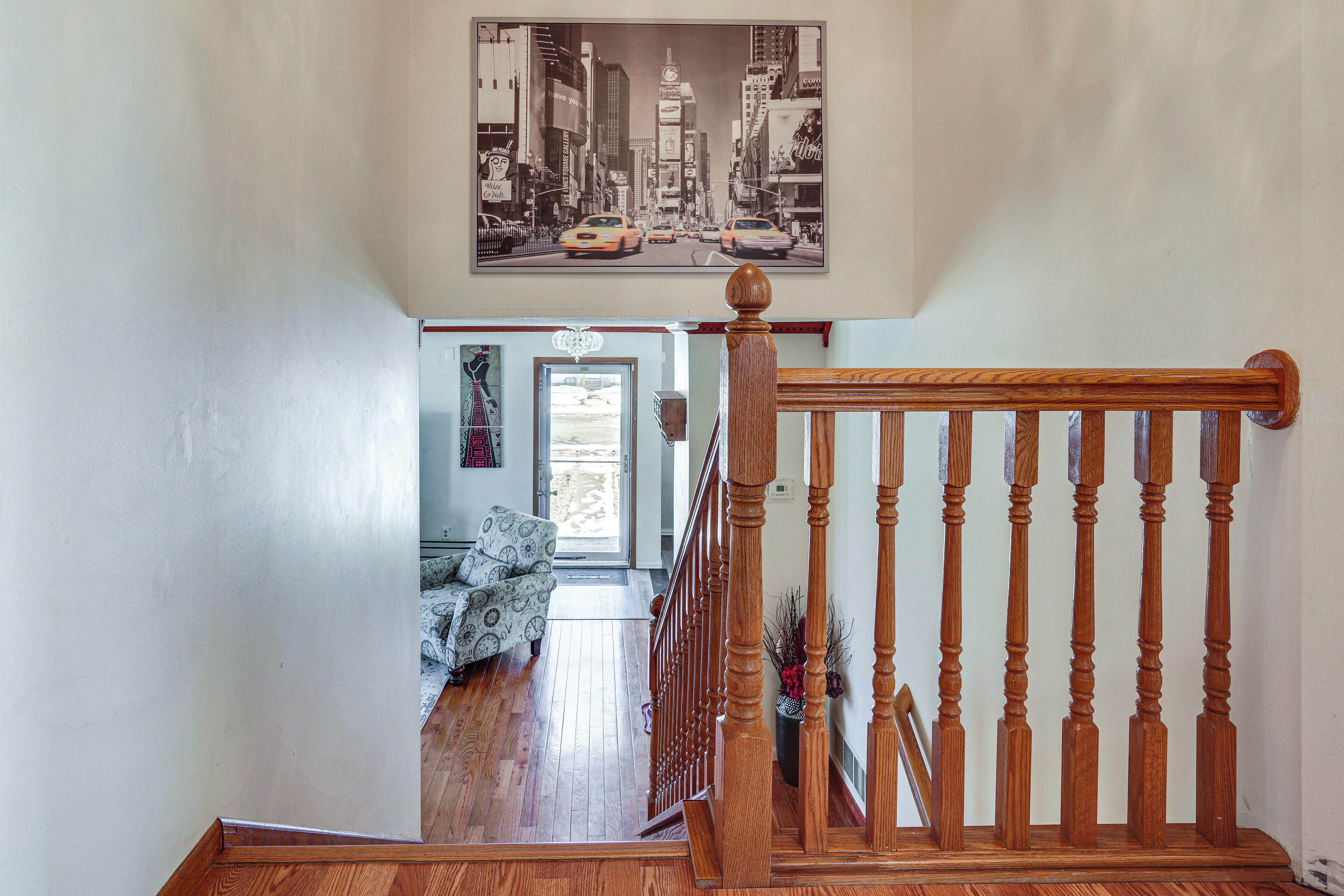 A staircase with a wooden railing leading to a living room with patterned furniture and a cityscape painting above.