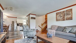 A spacious living room with a grey sofa, patterned armchair, wooden coffee table, and a chandelier. A staircase with wooden railings leads to an upper level.