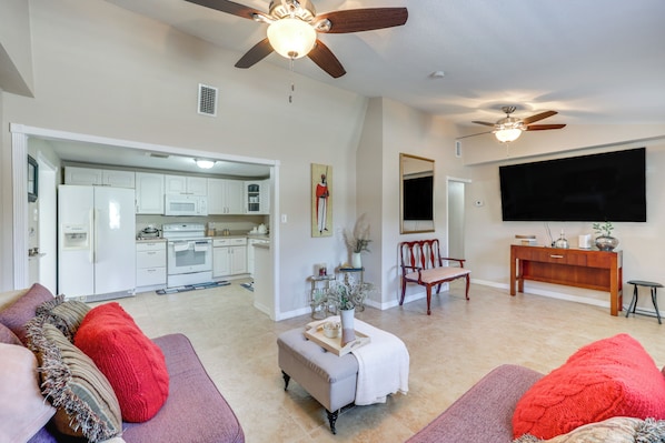 A spacious living room with a large TV, ceiling fans, and a kitchen in the background.