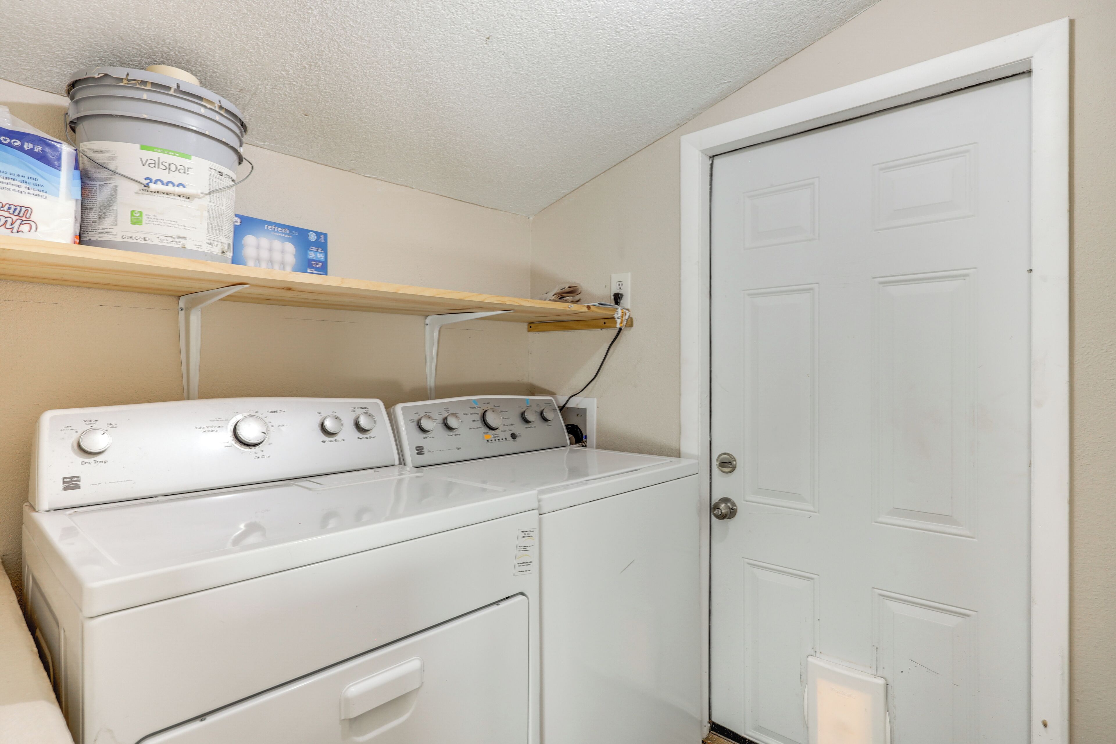A laundry area with a front-loading washer and dryer, a shelf with cleaning supplies, and a white door.