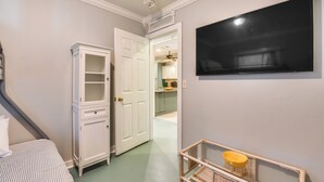 Un couloir avec une porte blanche donnant sur une cuisine, un téléviseur à écran plat fixé au mur et une chaise en osier avec un bol jaune.