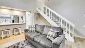 A modern living room with a gray sofa, two white bar stools, a kitchen area with a white countertop, and a white staircase. - Updated Tallahassee Townhome: 3 Mi to Downtown! (Tallahassee)