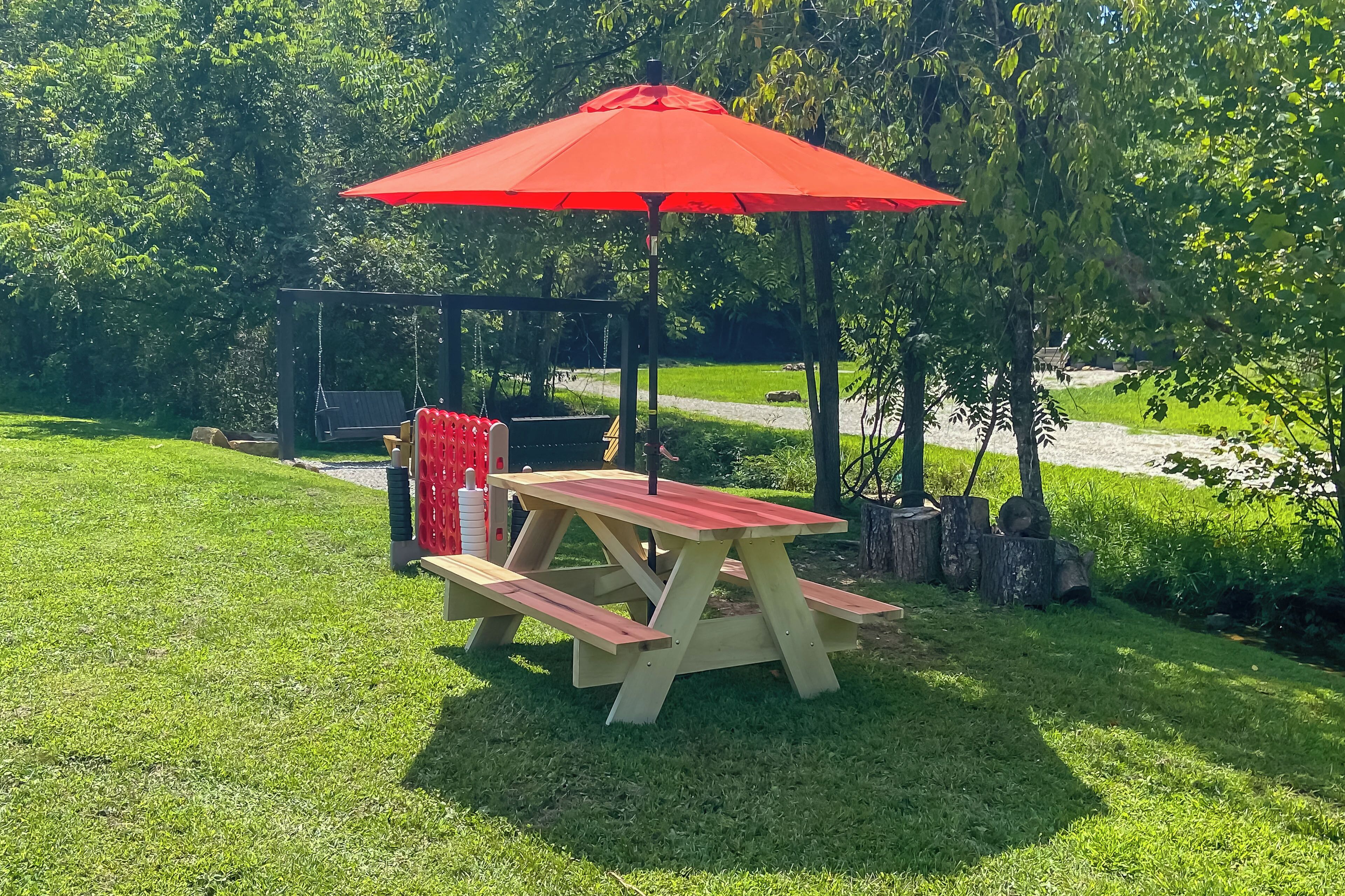A picnic table with a red umbrella, surrounded by a grassy area and trees.