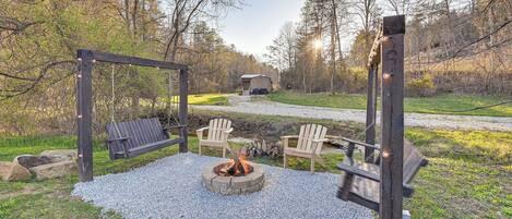 A fire pit surrounded by a wooden swing set and Adirondack chairs in a wooded outdoor area.