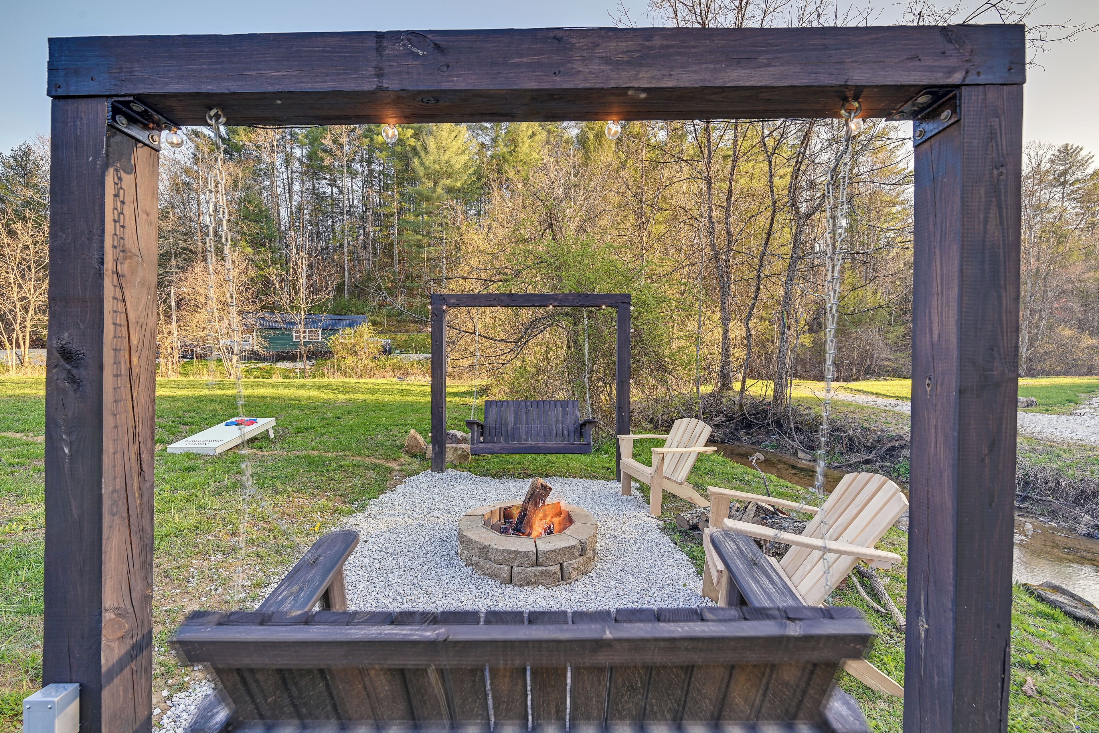 A wooden pergola with a fire pit in the center, surrounded by outdoor seating and a grassy area.