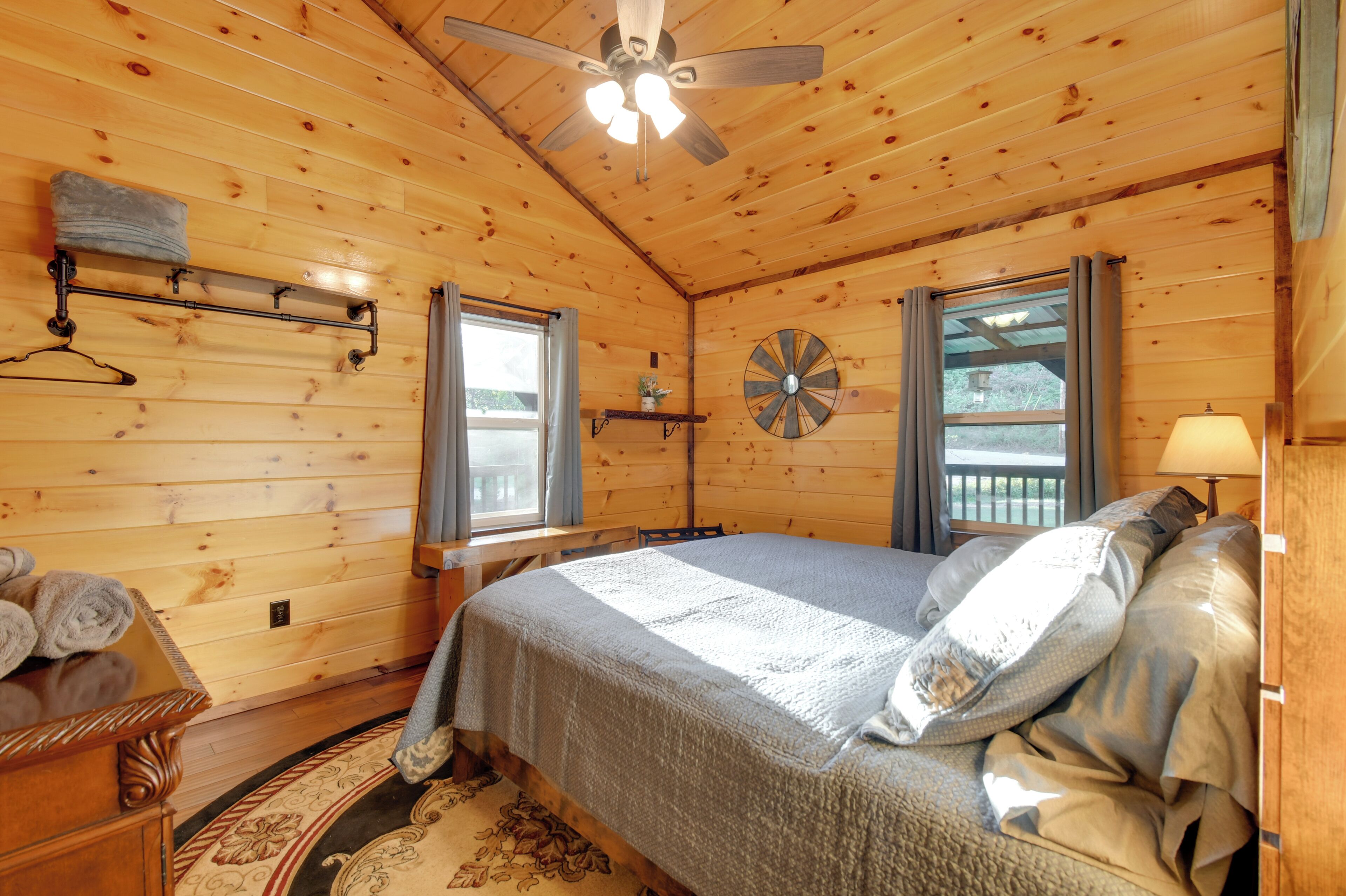 Un dormitorio de cabaña de madera con una cama, un ventilador de techo, una ventana, un reloj de pared y una lámpara.