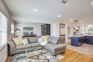 A modern living room with a sectional sofa, a glass coffee table, and a kitchen area with a blue island and stainless steel appliances.