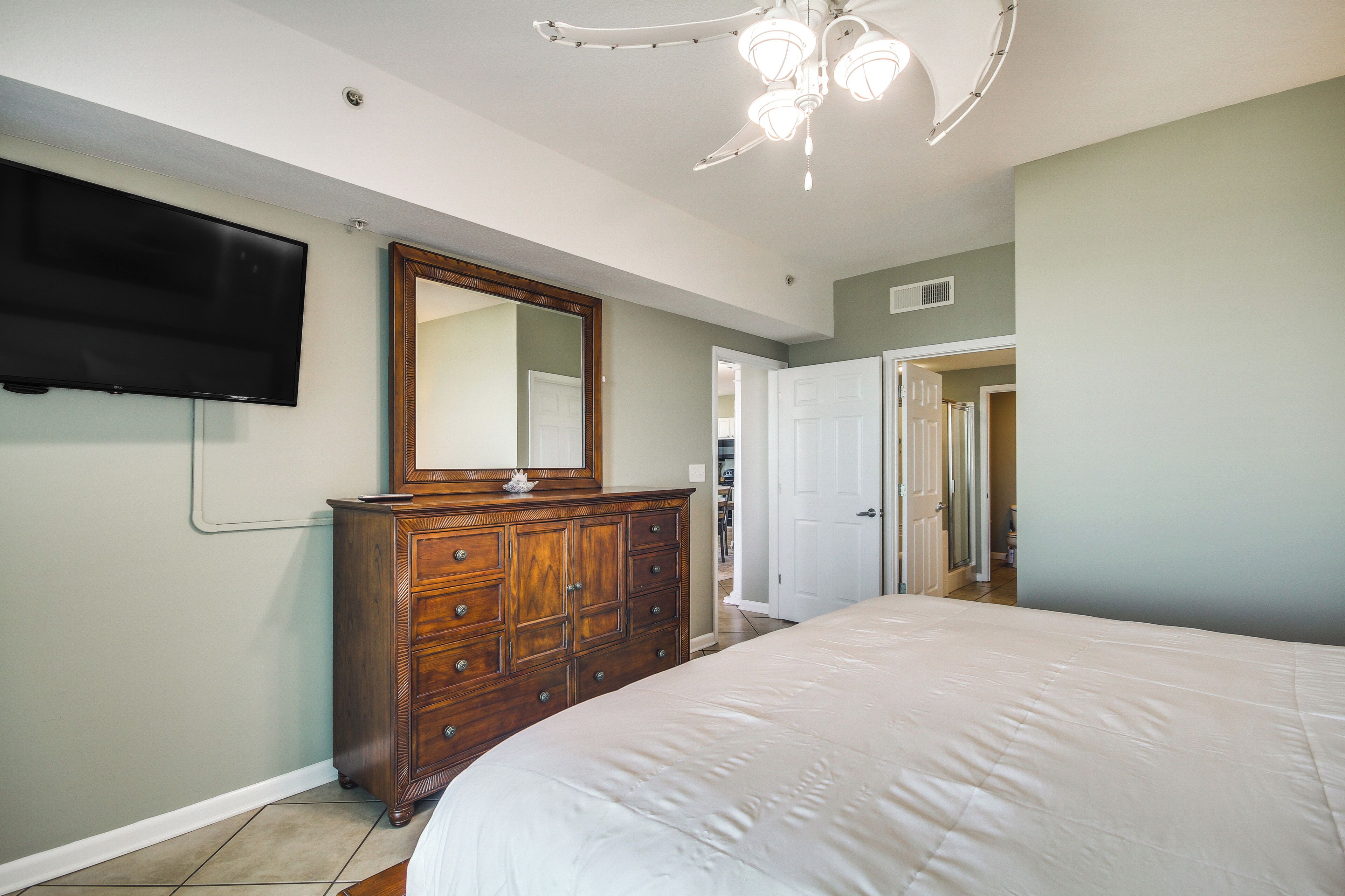 A bedroom with a large bed, a wooden dresser, a flat-screen TV, and a ceiling fan.