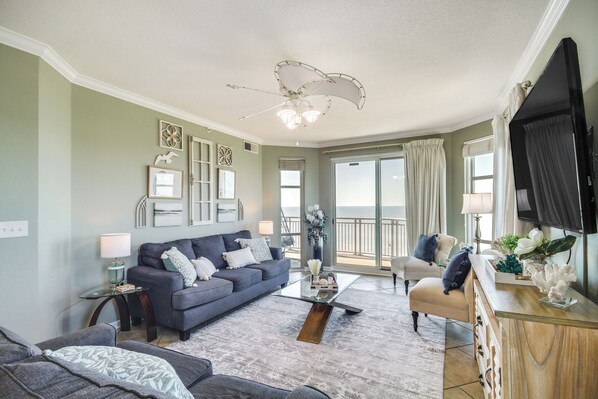 A living room with a blue sofa, beige armchair, and a glass coffee table. There is a TV mounted on the wall and a ceiling fan. - Gulfport Oceanfront Vacation Rental: Walk to Beach (Gulfport)