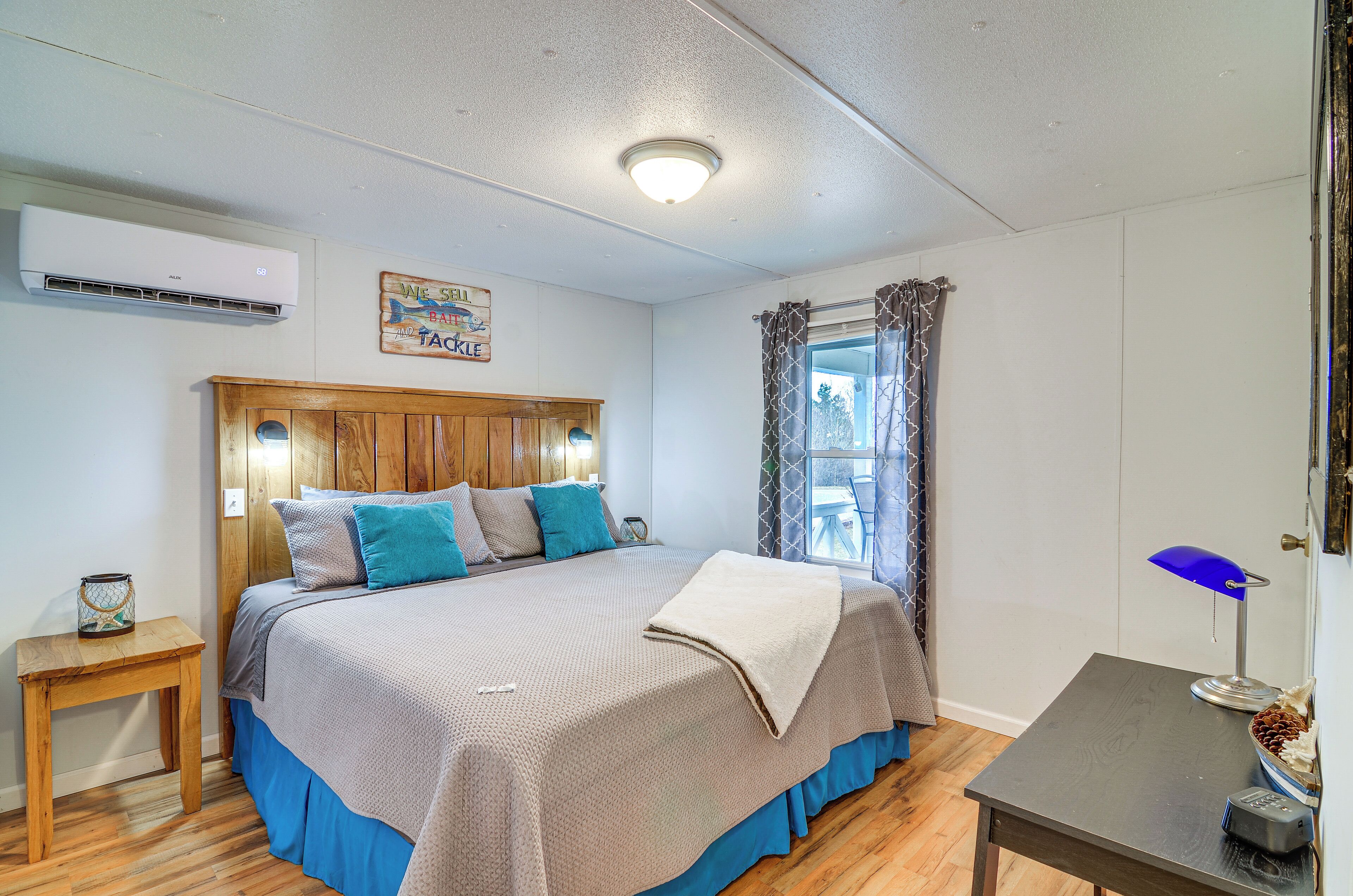 Un dormitorio con cabecera de madera, cama con ropa de cama azul y gris, una mesita de noche con lámpara y una ventana con cortinas.