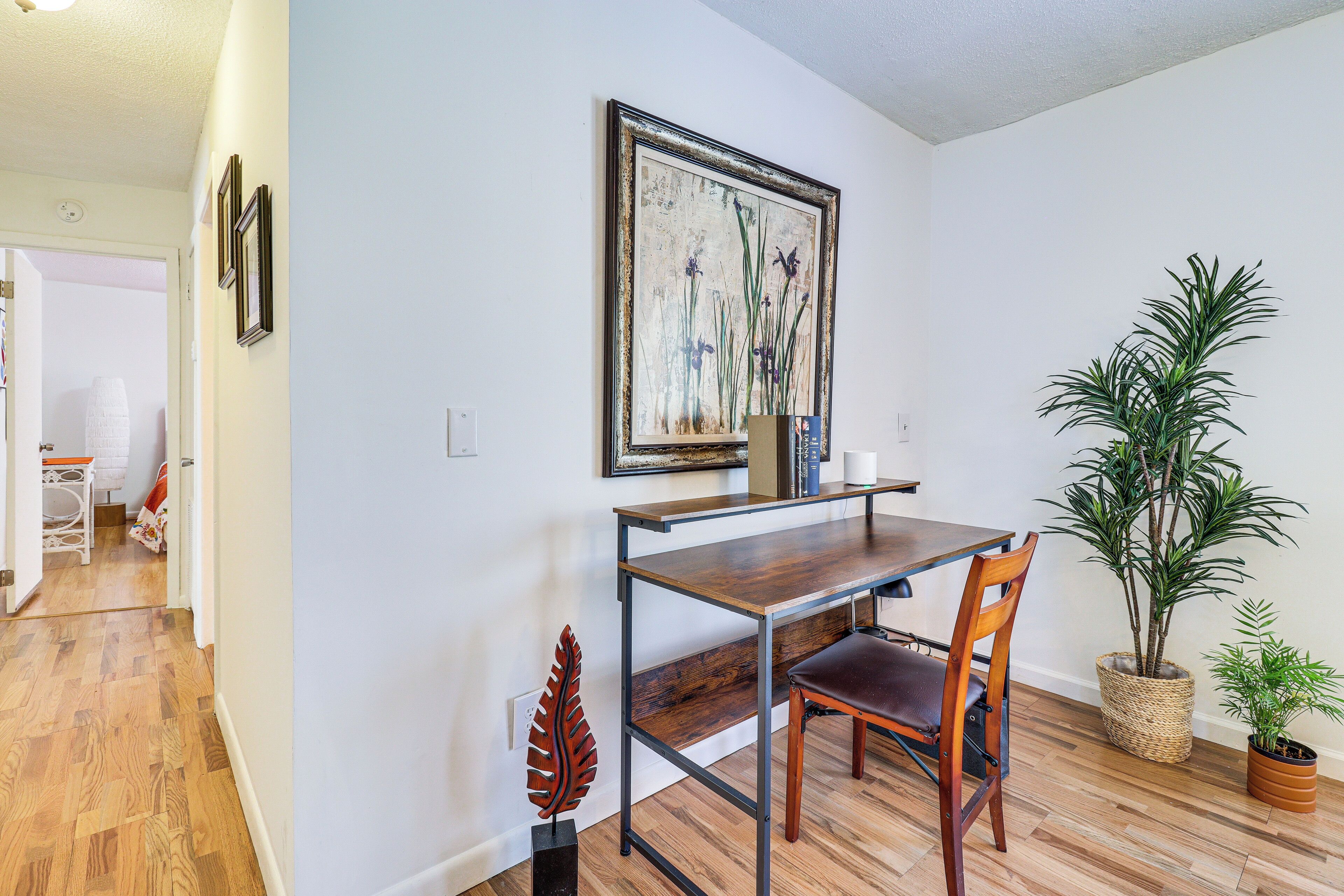 A dining area with a wooden table, two chairs, a framed painting, and two potted plants.