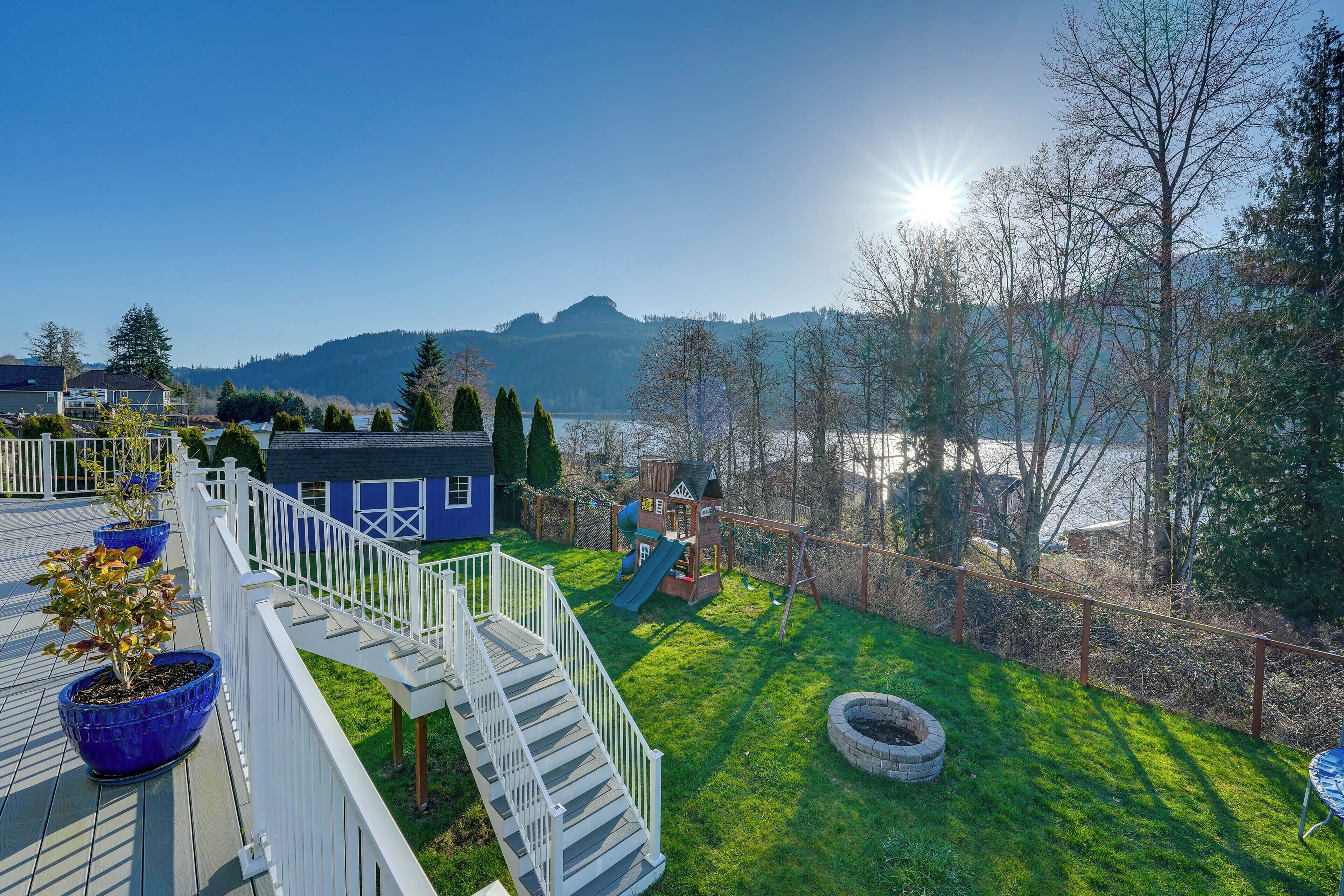 A residential backyard with a wooden deck, a playhouse, a swing set, and a view of a lake and mountains.