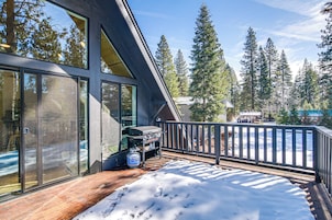 Una terraza de madera con nieve, una parrilla y vistas a altos árboles.