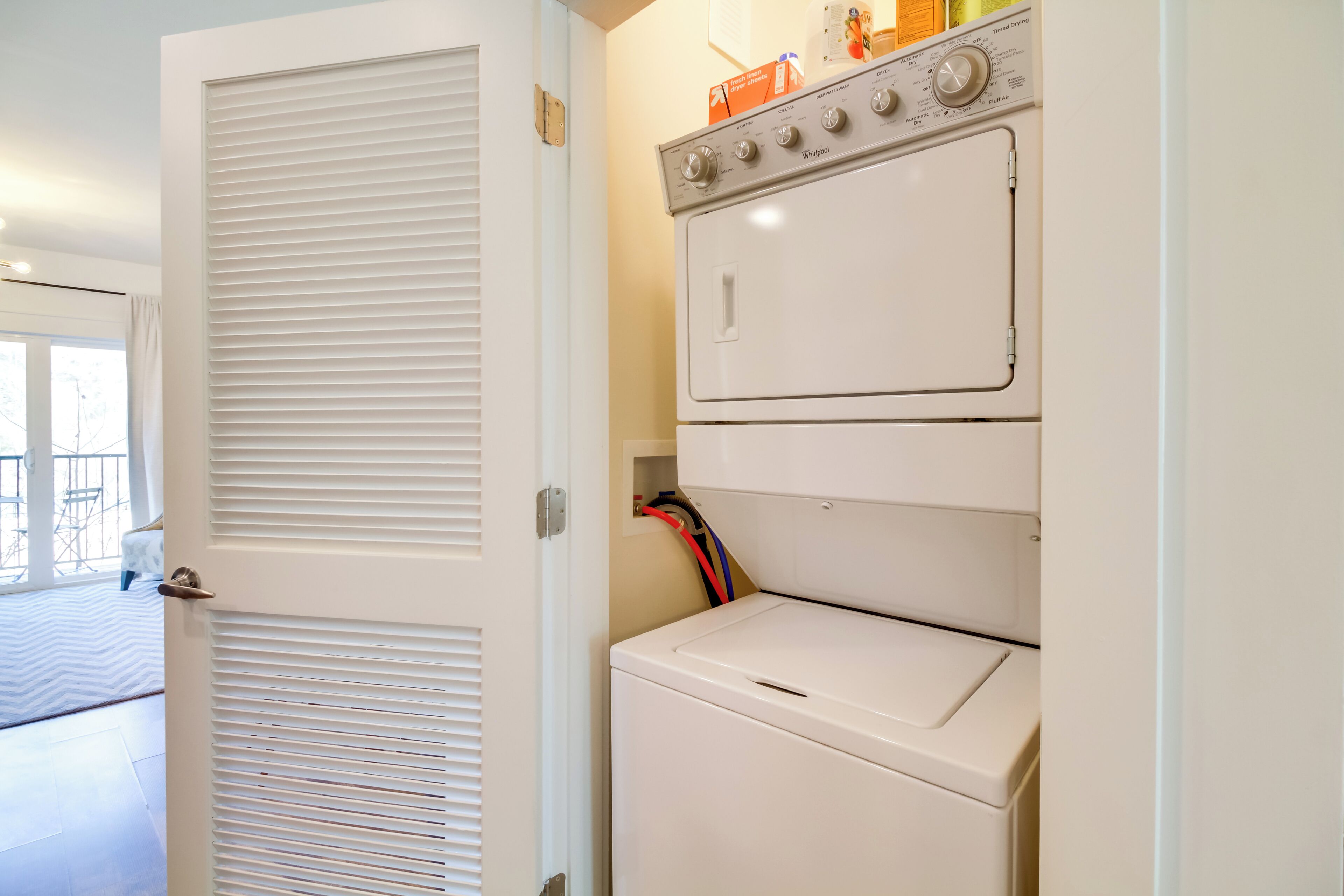 A laundry area with a stacked washer and dryer, a door with louvered panels, and a visible electrical outlet.